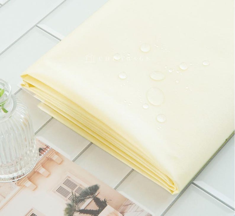 May include: A stack of folded, pale yellow fabric with water droplets on the surface. The fabric is neatly folded, revealing multiple layers. The background features a white tiled surface and a glass vase.