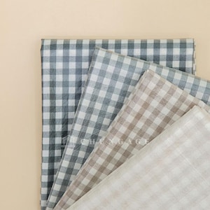 May include: A collection of folded fabric pieces in various colors, including gray, beige, and off-white. Each piece features a classic gingham check pattern. The fabrics are arranged on a light beige background.