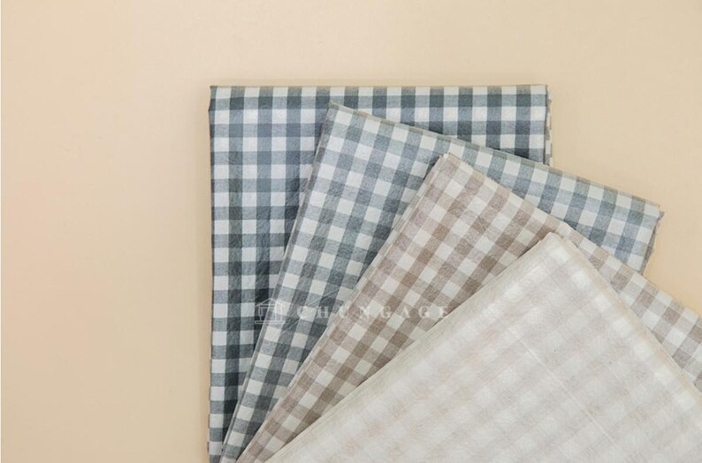 May include: A collection of folded fabric pieces in various colors, including gray, beige, and off-white. Each piece features a classic gingham check pattern. The fabrics are arranged on a light beige background.
