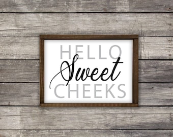 May include: A framed sign with the text "HELLO sweet CHEEKS" in black lettering on a white background. The frame is brown wood.