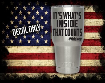 May include: A stainless steel tumbler with a black decal that reads "It's what's inside that counts - Whiskey". The tumbler is on a background of an American flag.