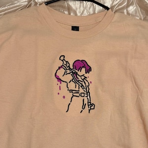 May include: A peach-colored crew neck t-shirt featuring an embroidered anime-style character with purple hair and accents. The character is holding a sword over their shoulder, with purple drips. The shirt is hanging on a black hanger.