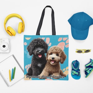 May include: A tote bag with a blue background featuring a black and brown doodle dog illustration. The text "Dynamic duo of doodles" is printed on the bag. Other items include yellow headphones, a white camera, a blue cap, sunglasses, sandals, and a notepad with crayons.