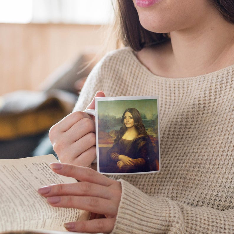 May include: A white ceramic mug with a colorful image of a woman with long brown hair, wearing a black dress, and a landscape background. The mug is being held by a person.