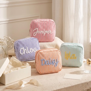 May include: Four square fabric pouches with vertical stripes in red, purple, orange, and light green. Each pouch features a white or yellow embroidered name in cursive script: Juniper, Chloe, Daisy, and Isla. Each has a metal zipper closure.