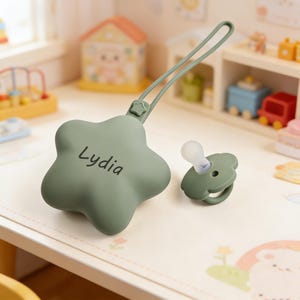 May include: A sage green silicone pacifier case shaped like a five-pointed star with the name Lydia printed in black, featuring a matching silicone strap and a coordinating sage green pacifier with a clear silicone nipple and a flower-shaped shield.