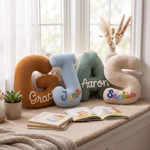 May include: Four plush, textured letter-shaped pillows in mustard, light blue, sage green, and cream. Each pillow features a personalized name embroidered in cursive script: Grace, June, Aaron, and Sophia. The letters are arranged in a row.