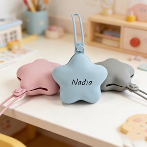 May include: Three star-shaped pacifier holders in pastel colors: pink, blue, and gray. The blue holder has the name "Nadia" printed on it. Each holder has a loop and a star-shaped clip.