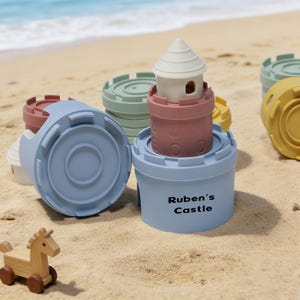 Könnte beinhalten: Ein Set aus stapelbaren Burgspielzeugen in Pastellfarben, darunter blaue, rosa, grüne und gelbe Teile, mit einem Holzpferdspielzeug im Sand. Auf einem der blauen Burgteile steht der Text "Ruben's Castle".