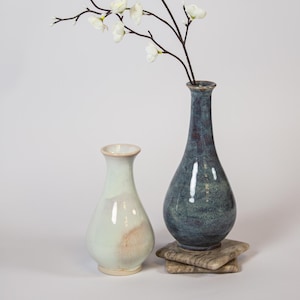 May include: Two ceramic vases with narrow necks and rounded bodies. One vase is a pale, glossy off-white with subtle tan accents, while the other is a deep, mottled blue-grey. A single branch with small white flowers extends from the blue vase.