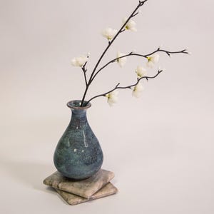 May include: A small, bulbous ceramic vase with a mottled blue and green glaze. The vase holds a slender, dark brown branch featuring several small, white, five-petaled blossoms and tiny buds, extending outward in a natural, branching pattern.