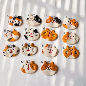 May include: A collection of fourteen ceramic cat-shaped pendants labeled A through N. Each pendant features a unique hand-painted face with whiskers, a small nose, and a metal loop at the top for hanging. The cats are white with orange and black spots.