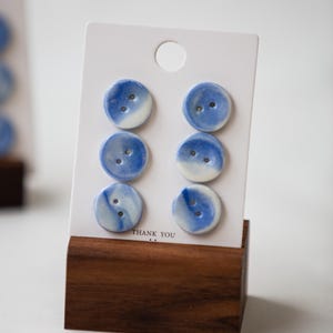 May include: Six round ceramic buttons with a blue and white marbled pattern, each featuring two central holes. The buttons are arranged in two vertical columns of three on a white rectangular display card with the text THANK YOU printed at the bottom.