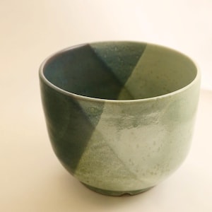 May include: A handmade ceramic bowl with a unique design. The bowl features a geometric pattern with shades of green. The interior and exterior surfaces have a matte finish. The bowl is suitable for serving food or as a decorative piece.