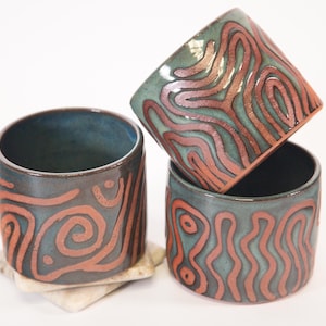 May include: Three handmade ceramic cups with a teal glaze and a reddish-brown, abstract, linear design. One cup is stacked on top of the other two. The cups are cylindrical and rest on a white surface.