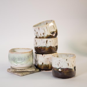 May include: A collection of five ceramic cups with a smooth, rounded shape. Three cups are stacked vertically, one stands alone, and one rests on a square stone coaster. The glaze features a white upper section with brown drips and a dark brown base.