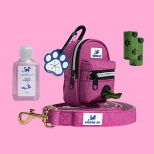 Op de afbeelding: Een roze hondenwandelset met een kleine rugzak, een riem, een tag in de vorm van een poot, een flesje handdesinfectie en twee rollen poepzakjes. De rugzak heeft de tekst "POOPSIE KIT" erop. De riem heeft ook de tekst "POOPSIE KIT" erop.