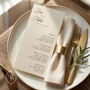 Puede incluir: Una tarjeta de menú de boda color crema con los nombres Bea y Nate y la fecha 05.10.2027, colocada sobre un plato moteado junto a una servilleta de lino beige con un anillo dorado, un tenedor dorado y un cuchillo dorado con una rama.