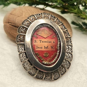 Pode incluir: Relicário antigo prateado com um centro oval. O centro apresenta um fundo de tecido vermelho com o texto "S. Teresiae a Jesu Inf. V." O relicário tem detalhes intrincados de filigrana nas bordas.