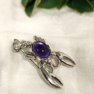 May include: A silver-toned lobster brooch with a large, oval, purple gemstone at its center. The brooch has detailed claws and a pin backing. Green leaves are blurred in the background. A unique jewelry piece.
