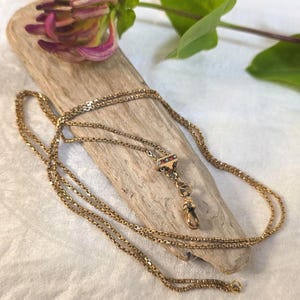 May include: A gold-toned metal chain necklace featuring a delicate link design. The necklace includes a small, rectangular decorative element set with tiny red and white stones, and a teardrop-shaped pendant clasp hanging from the centre.