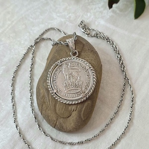 Peut inclure: Un collier argenté avec un pendentif circulaire fabriqué à partir d'une pièce de monnaie britannique d'un shilling de 1934. La pièce représente un lion debout sur une couronne avec le texte ONE SHILLING et FID DEF IND IMP sur une chaîne torsadée.