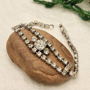 May include: A silver bracelet adorned with clear rhinestones. The design includes a central, large round stone, with two parallel rows of smaller stones. The bracelet is presented on a piece of wood.