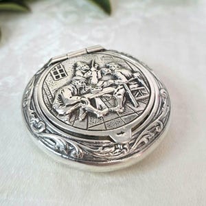 Puede incluir: Un estuche compacto redondo de metal plateado con una escena en relieve de tres figuras sentadas a una mesa en la tapa. El borde exterior de la tapa está decorado con un patrón floral en relieve y el estuche tiene un mecanismo de cierre.
