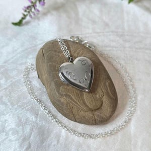 May include: A silver-toned heart-shaped locket pendant suspended from a delicate matching silver-toned chain. The locket features an engraved floral pattern on its front surface, showcasing a polished metallic finish and a classic hinged design.