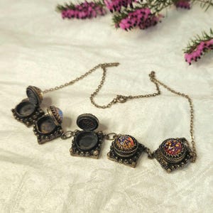 May include: A vintage-style necklace with four ornate, hinged compartments. The compartments are bronze-toned with intricate detailing. Two of the compartments are open, revealing a small interior space. The necklace features colorful, mosaic-like glass cabochons.