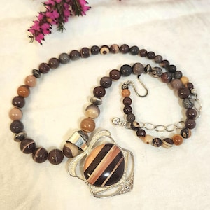 May include: A beaded necklace with a large pendant. The necklace features brown, tan, and gray beads. The pendant has a silver-colored frame and a striped, oval-shaped center in shades of brown and tan.