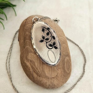 Peut inclure: Pendentif ovale en argent avec un motif floral et une chaîne délicate. Le médaillon présente un motif floral noir sur un fond rayé. La chaîne est de style corde fine, de couleur argent.