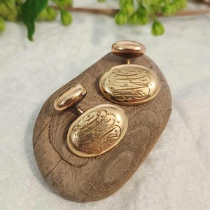 Könnte beinhalten: Goldfarbene Manschettenknöpfe mit graviertem Design. Die Manschettenknöpfe sind oval und kreisförmig und liegen auf einem Holzstück. Der Hintergrund umfasst Grün und weißen Stoff. Diese Accessoires eignen sich für formelle Anlässe.
