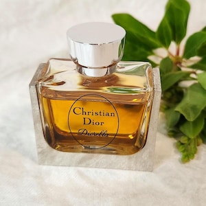 May include: A square perfume bottle with a silver cap and a clear glass body. The bottle contains a golden-colored liquid and features the text "Christian Dior Diorella" on a circular label. Green leaves are in the background.