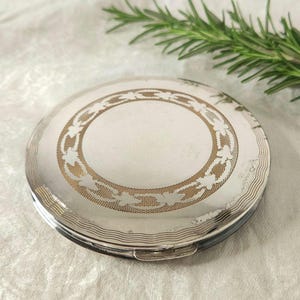 Op de afbeelding: Een zilverkleurige compacte poederdoos met een decoratief goudkleurig bloemmotief in het midden. De ronde compacte poederdoos heeft een scharnierende opening. Een takje rozemarijn staat op de achtergrond.