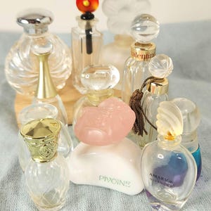 May include: A selection of miniature perfume bottles in diverse shapes and styles. Some are clear glass, others have colourful details and distinctive stoppers. The bottles are displayed on a light blue surface.