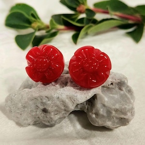 Puede incluir: Dos pendientes de botón rojos brillantes con forma de flor. Los pendientes, hechos de un material brillante, descansan sobre una piedra gris. Hojas verdes en el fondo. Un accesorio elegante.