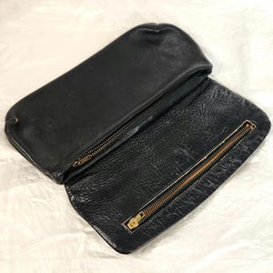Könnte beinhalten: Schwarze Leder-Clutch mit rechteckiger Form, geöffnet, um das Innere zu zeigen. Die Clutch hat einen Messing-Reißverschluss und eine strukturierte Lederoberfläche. Das offene Design zeigt das Innenfutter und die Reißverschlussdetails.