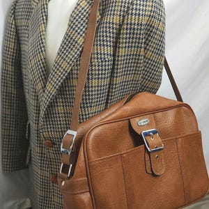 Può includere: Una borsa da viaggio in pelle marrone con una fibbia argentata e una tracolla. La borsa ha il testo "Jet Stream" sul davanti.