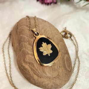 May include: A gold-coloured pendant necklace with a black enamel inlay and a gold maple leaf design. The teardrop-shaped pendant hangs from a delicate chain. The necklace is displayed on a piece of driftwood.