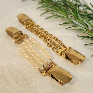 Puede incluir: Dos pinzas de tirantes vintage de metal dorado. Una pinza presenta tres hilos de perlas de imitación, mientras que la otra tiene un diseño de doble cadena. Las pinzas están sobre una superficie blanca con una ramita de romero.