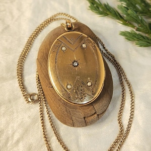Puede incluir: Un collar con colgante de medallón ovalado dorado con un diseño detallado. El medallón presenta un diseño central en forma de estrella con una pequeña gema, rodeado de elementos decorativos y pequeñas perlas. La cadena del collar también es dorada.