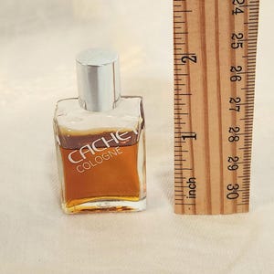 May include: A small, square glass bottle of "Cache Cologne" with a silver cap. The bottle contains a golden-brown liquid. A wooden ruler is next to the bottle, showing measurements in inches.