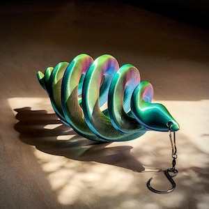 May include: A vibrant, iridescent sculpture in the shape of a coiled snake. The sculpture transitions through shades of green, blue, and purple. A small hook hangs from the snake's head. The sculpture casts a shadow on a light-colored surface.