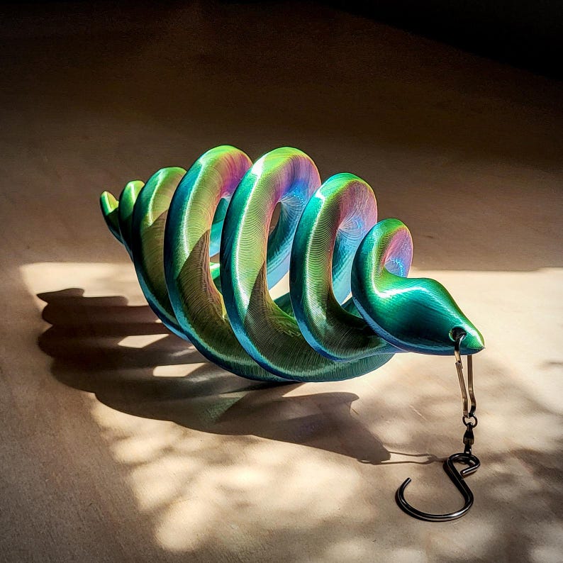 May include: A vibrant, iridescent sculpture in the shape of a coiled snake. The sculpture transitions through shades of green, blue, and purple. A small hook hangs from the snake's head. The sculpture casts a shadow on a light-colored surface.