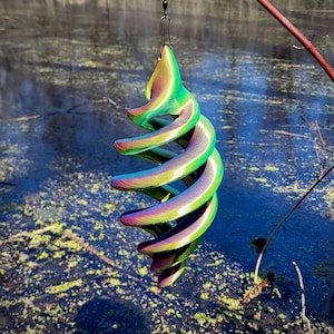 May include: A spiral-shaped wind spinner with a metallic, iridescent finish. The colors shift from green to purple and gold. The decorative item is suspended by a black hook, with a blurred water background.