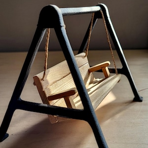 Könnte beinhalten: Ein Miniatur-Holzschaukelgestell mit Sitz und Rückenlehne aus Naturholz, das an Seilen von einem schwarzen Metallrahmen abgehängt ist. Die Schaukel hat ein schlichtes Design und steht vor einem neutralen Hintergrund.