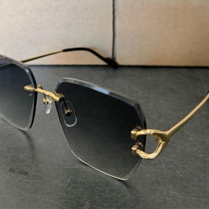 May include: Rimless sunglasses with a gradient lens. The frames are gold-coloured with a distinctive design at the temples. The lenses transition from dark at the top to clear at the bottom. The sunglasses are set against a neutral background.