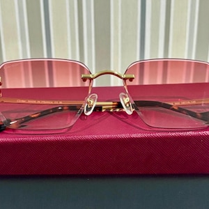 May include: Rimless sunglasses with pink tinted lenses and gold-colored metal frames. The temples have a tortoiseshell pattern and black tips. The glasses are resting on a red box.