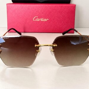 May include: Rimless sunglasses with gradient brown lenses and gold-tone metal temples. The Cartier logo is printed on the upper corner of the left lens. The glasses are positioned in front of a rectangular red box with the Cartier logo embossed.
